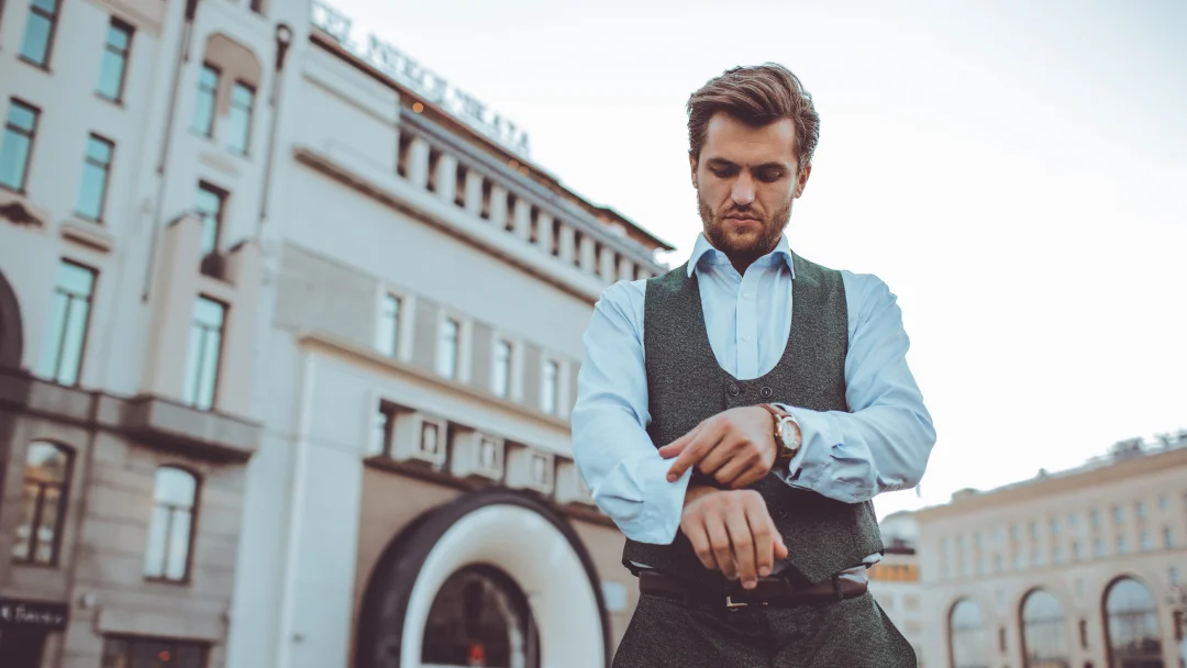 Un homme en gilet et chemise regarde sa montre, semblant impatient, dans un contexte de nouveautés.