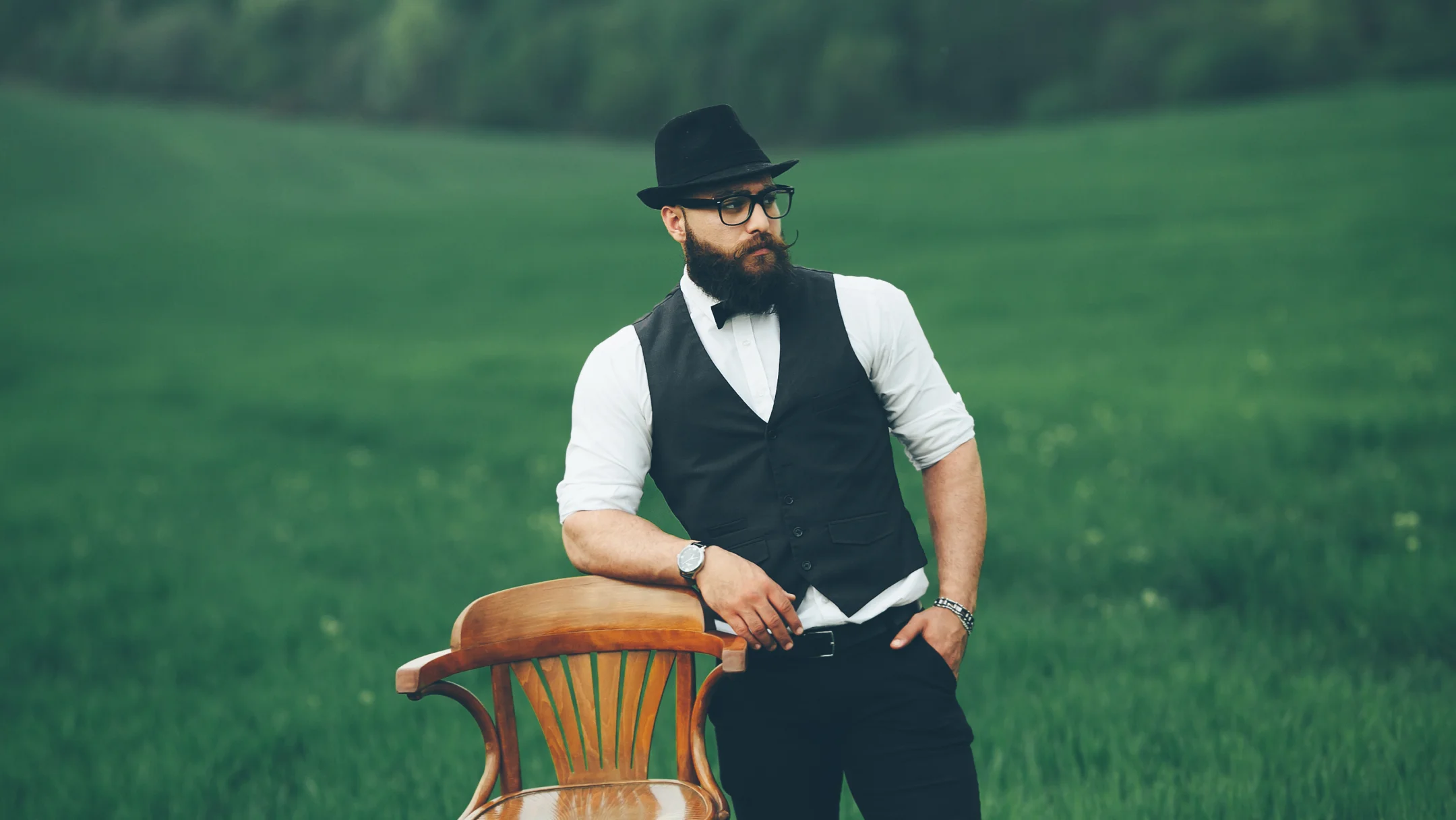 Un homme barbu et portant des lunettes, habillé comme dans Peaky Blinders, se tient dans un champ verdoyant.