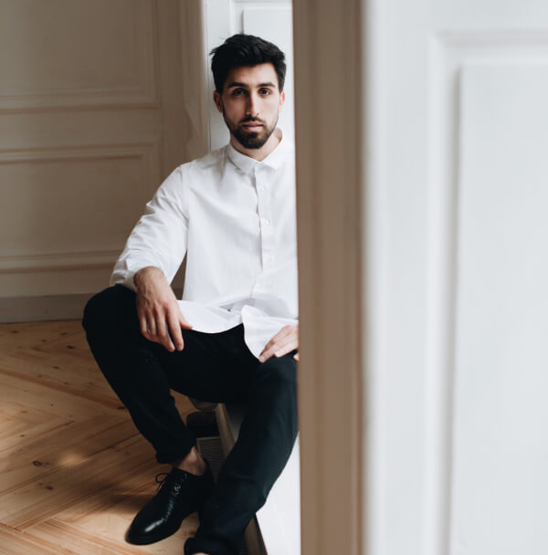 Un homme en chemise blanche et chaussures noires assis sur un sol en bois, illustrant l'élégance masculine.
