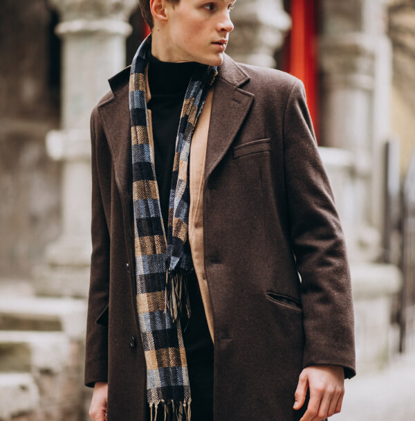 Un homme en manteau marron et écharpe se tient dans la rue, illustrant la tendance masculine de 2026 : retour à l’essentiel.