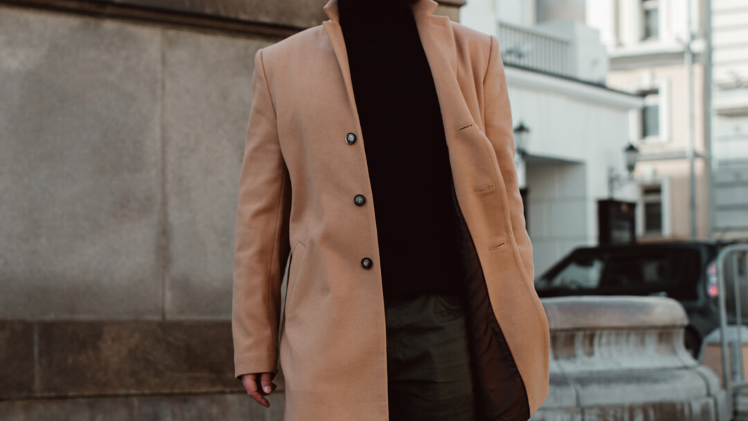 Un homme portant un manteau camel et un col roulé noir, se tenant debout avec un air élégant.