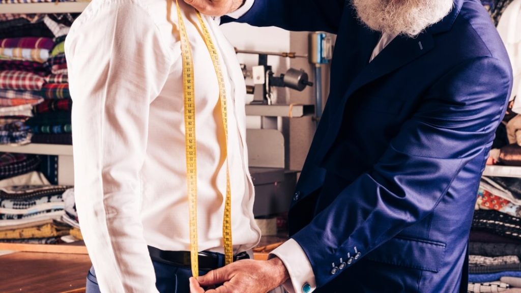 Une vue perspective d'une boutique ou d'un atelier de haute couture pour hommes à Paris, montrant des costumes sophistiqués sur des mannequins, des rouleaux de tissus fins et un décor classique raffiné.