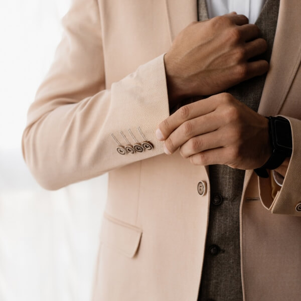 Une vue de trois quarts d'un homme souriant et bien habillé, ajustant le bouton de sa veste de costume haut de gamme en souriant, illustrant la confiance et le confort d'une coupe sur-mesure lors d'un événement spécial.