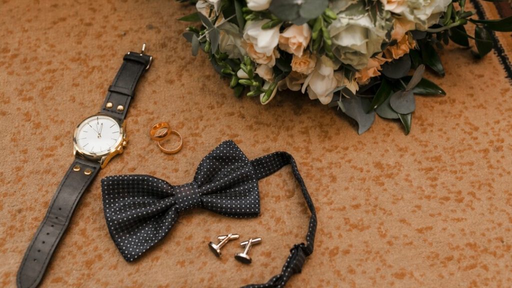 Une composition soignée d'accessoires de mariage pour homme disposés sur une surface élégante, incluant des boutons de manchette, une montre en cuir, une pochette de costume pliée et une paire de chaussures en cuir verni.